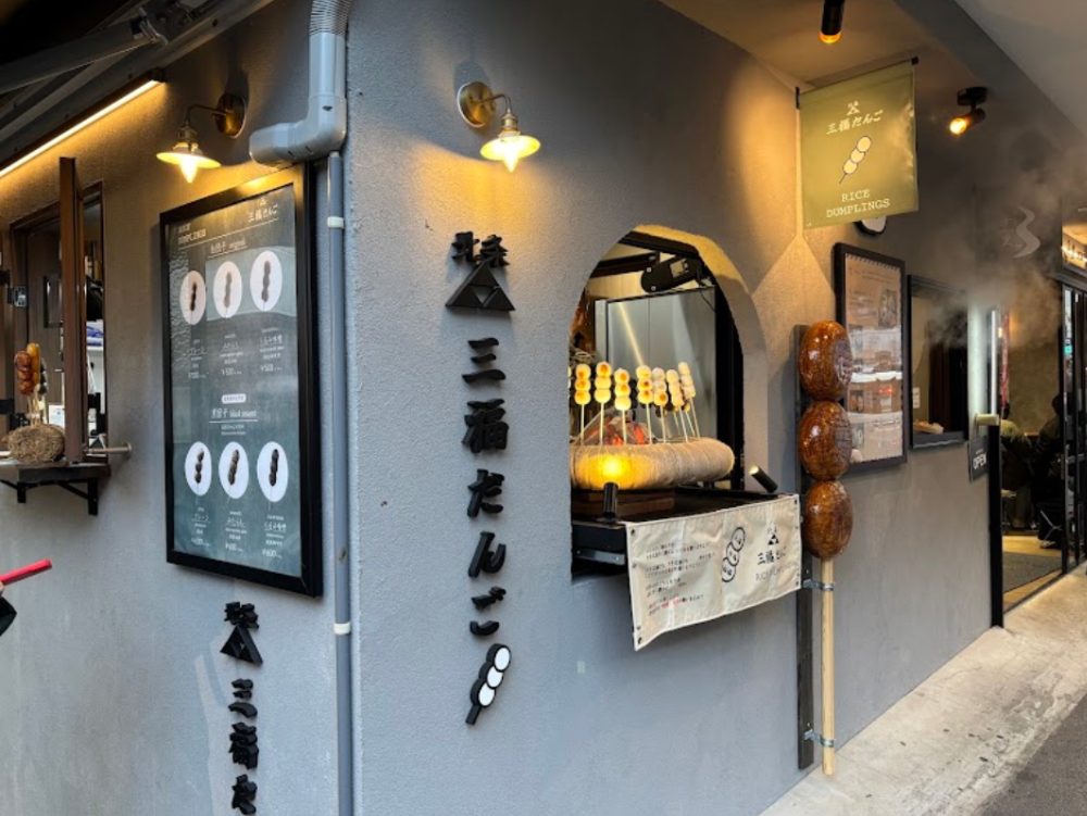 北条三福だんごの店構えの写真。灰色の壁に縦書きの店名、品書きの看板、串だんごの窓口